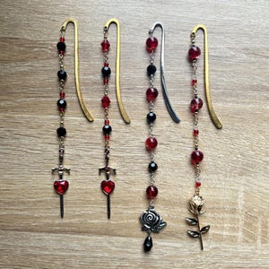 May include: Five decorative bookmarks with dangling charms. The bookmarks feature red and black beads, with charms including a sword with a heart, a rose, and a gold rose. The bookmarks are on a wooden surface.