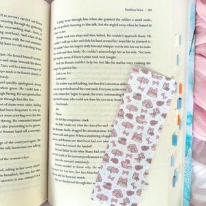 Peut inclure: Marque-page décoratif avec un motif répété de nœuds roses, de gâteaux et de roses. Le marque-page est rectangulaire avec des coins arrondis et est posé sur les pages d'un livre ouvert.