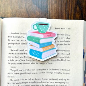 Könnte beinhalten: Ein farbenfroher Lesezeichen mit einem Stapel Bücher und einer Kaffeetasse. Die Bücher sind in Rosa, Grün, Blau und Orange gehalten. Die Kaffeetasse ist türkis mit weißen Punkten. Das Lesezeichen befindet sich in einem offenen Buch.