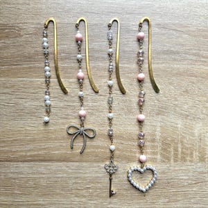 May include: Four decorative gold-tone metal bookmarks with dangling beaded accents. The beads are white, pink, and clear, with charms including a bow, a key, and a heart. The bookmarks are arranged on a light brown wooden surface.