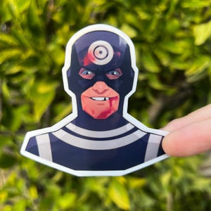 May include: A sticker featuring a comic book character with a target symbol on the forehead. The character has a dark blue mask, a red mouth, and blue eyes. The sticker is held by a hand against a blurred green background.