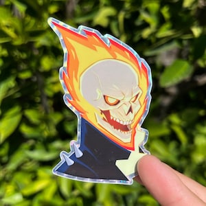 May include: Holographic sticker featuring the Ghost Rider character. The sticker depicts a flaming skull with orange and yellow flames, a black jacket, and silver spikes. The sticker has a shiny, reflective surface.