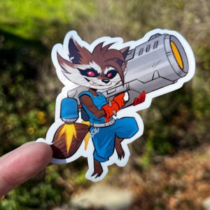 May include: A sticker featuring a cartoon raccoon character in a blue outfit, holding a large silver weapon with an orange barrel. The raccoon has brown fur, red eyes, and is wearing orange gloves. The sticker is held up by a finger.