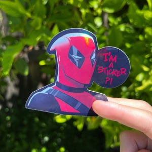 May include: A sticker depicting Deadpool's head in red and black, with a speech bubble saying "I'M A STICKER ?!". The sticker is held against a blurred green backdrop.