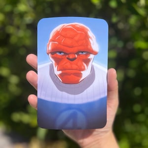 May include: A rectangular card featuring a stylized illustration of a red-skinned character with a stern expression and blue eyes. The character is wearing a white shirt with a blue collar, set against a blue background.