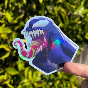 May include: Holographic sticker featuring a stylized illustration of Venom, a comic book character. The sticker has a dark blue background with a white face, sharp teeth, and a long, pink and green tongue. The sticker is held against a blurred green background.