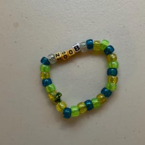 Könnte beinhalten: Ein kreisförmiges Armband aus durchscheinenden Kunststoffperlen in abwechselnden Farbtönen von Limettengrün und dunklem Blaugrün. Das Armband enthält vier quadratische weiße Perlen mit schwarzer Beschriftung, die das Wort NOOB bilden, durchsetzt mit klaren Perlen.
