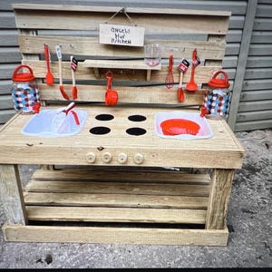 May include: A wooden play kitchen with a red and white checkered container, red plastic utensils, and a sign that reads "Angels Mud Kitchen".