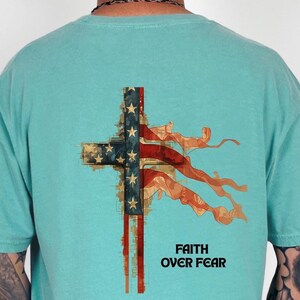 Può includere: T-shirt verde acqua chiaro con un design a croce che incorpora la bandiera americana. La croce presenta stelle e strisce, con la scritta "FAITH OVER FEAR" stampata sotto. La maglietta è realizzata in materiale morbido.
