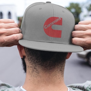 Peut inclure: Casquette de baseball grise à visière plate avec le logo rouge "Cummins". La casquette est tenue par une personne. La personne porte une chemise à manches longues à motifs verts et blancs.