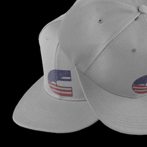 Peut inclure: Deux casquettes de baseball grises avec un motif de drapeau américain rouge, blanc et bleu sur le devant. Les casquettes sont toutes les deux à plat.