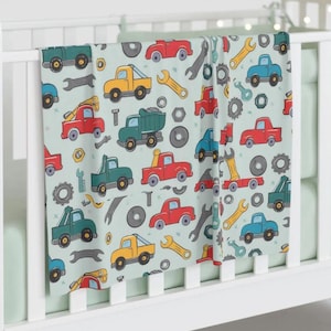 May include: A baby blanket with a light blue background and a repeating pattern of toy trucks, wrenches, and nuts and bolts. The trucks are red, yellow, green, and blue.