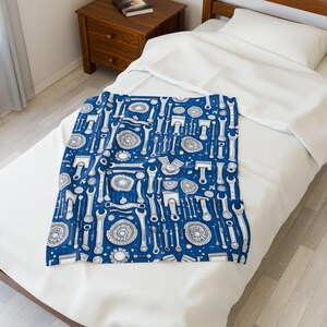 May include: A blue blanket with a repeating pattern of white tools, including wrenches, gears, and other mechanical components. The blanket is spread out on a white bed, with a wooden nightstand in the background.