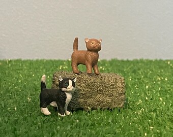 Könnte beinhalten: Zwei kleine Spielzeugkatzen, eine braun und eine schwarz-weiß, stehen auf einem kleinen Heuballen. Die braune Katze steht oben auf dem Heuballen, während die schwarz-weiße Katze davor steht.