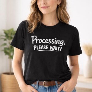 May include: Black crew neck t-shirt with the text "Processing. PLEASE WAIT!" in white script. The model is wearing blue jeans and a brown belt. The background includes a potted plant and a vase.