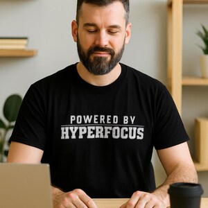 May include: Black t-shirt with the text "POWERED BY HYPERFOCUS" in white. A man with a beard is wearing the shirt and sitting at a desk with a laptop, notebook, and coffee cup. The background includes shelves.