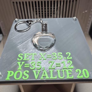 May include: A clear, heart-shaped keychain charm with a silver-coloured metal top is attached to a silver keyring. The charm sits on a grey platform with green text that reads "SET X=35.2 Y=35 Z=12 POS VALUE 20".