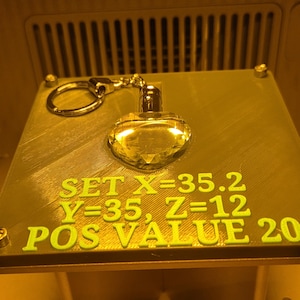 Op de afbeelding: Een goudkleurige hartvormige sleutelhanger met een facetontwerp. De sleutelhanger is bevestigd aan een zilverkleurige ring. De tekst "SET X=35.2 Y=35, Z=12 POS VALUE 20" is groen op het oppervlak gedrukt.