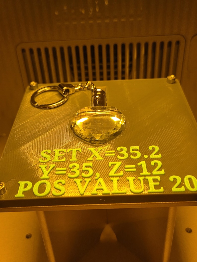 Op de afbeelding: Een goudkleurige hartvormige sleutelhanger met een facetontwerp. De sleutelhanger is bevestigd aan een zilverkleurige ring. De tekst "SET X=35.2 Y=35, Z=12 POS VALUE 20" is groen op het oppervlak gedrukt.