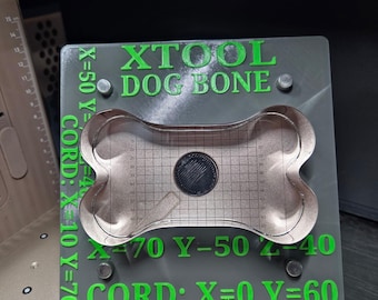 Könnte beinhalten: Eine graue quadratische Grundplatte mit grünem Text XTOOL DOG BONE und verschiedenen Koordinatenbeschriftungen. Ein metallischer, hundeknochenförmiger Einsatz mit aufgedrucktem Gittermuster ist mittig auf der Platte platziert und mit vier kleinen Befestigungen gesichert.