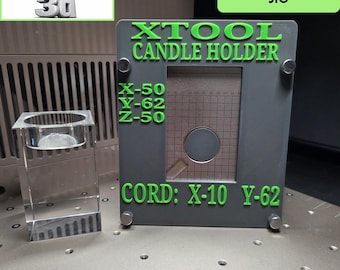 Könnte beinhalten: Eine graue, rechteckige 3D-gedruckte Vorrichtung mit grünem Text: XTOOL CANDLE HOLDER, X-50, Y-62, Z-50 und CORD: X-10 Y-62. Die Vorrichtung hat ein zentrales Rasterfenster und vier Eckschrauben und steht neben einem Kerzenhalter aus Glas.