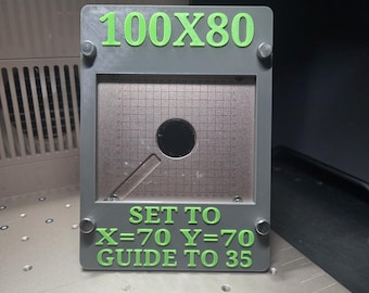 Könnte beinhalten: Ein rechteckiger grauer Rahmen mit hellgrünem Text, der 100X80, SET TO X=70 Y=70 und GUIDE TO 35 liest. Die Mitte verfügt über eine metallische Gitterplatte mit einem kreisförmigen Ausschnitt und einem diagonalen Schlitz, befestigt durch vier kleine silberne Metallverschlüsse.