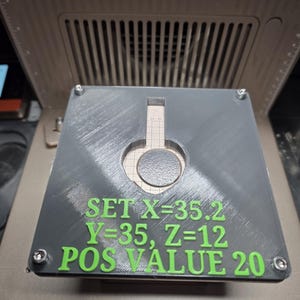 Op de afbeelding: Een grijs, vierkant object met een cirkelvormige uitsparing en groene tekst met de tekst "SET X=35.2 Y=35, Z=12 POS VALUE 20." Het object is bevestigd met vier schroeven en ligt op een metalen oppervlak.