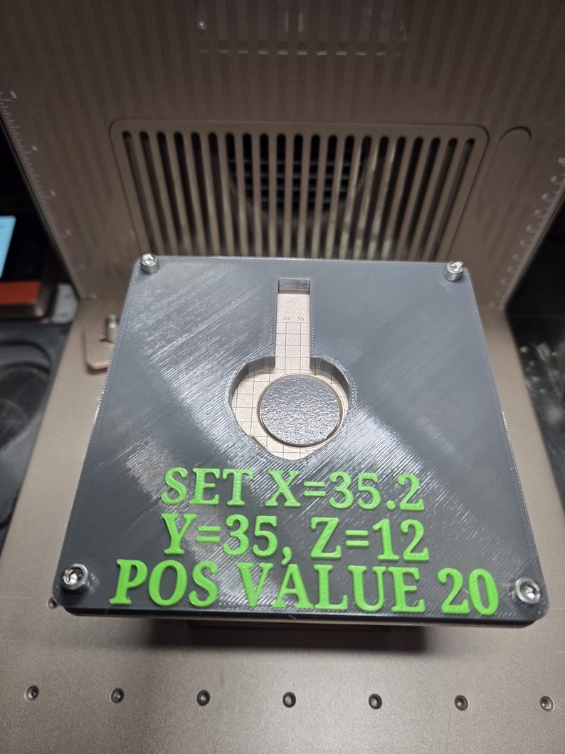 Op de afbeelding: Een grijs, vierkant object met een cirkelvormige uitsparing en groene tekst met de tekst "SET X=35.2 Y=35, Z=12 POS VALUE 20." Het object is bevestigd met vier schroeven en ligt op een metalen oppervlak.