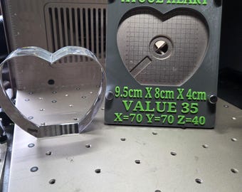 Op de afbeelding: Een helder glazen hartvormig object naast een grijze rechthoekige plaat met groene tekst die XTOOL HEART, 9.5cm X 8cm X 4cm, VALUE 35, X=70 Y=70 Z=40 leest. De plaat heeft een hartvormige uitsparing met een rasterpatroon en een cirkel.