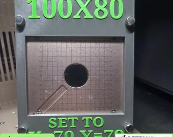 Könnte beinhalten: Ein graues, rechteckiges, 3D-gedrucktes Kalibrierungswerkzeug mit hellgrünem Text, der 100X80 und SET TO X=70 Y=70 liest. Die Mitte zeigt eine metallische Gitterplatte mit einem kreisförmigen Ausschnitt und einem diagonalen Balken.
