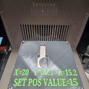 Könnte beinhalten: Ein graues, rechteckiges 3D-gedrucktes Objekt mit einem Ausschnitt und grünem Text mit der Aufschrift "X=20 Y=30.3 Z=15.2 SET POS VALUE=15." Das Objekt ist mit Schrauben befestigt und liegt auf einer Maschine mit Lüftungsschlitzen.