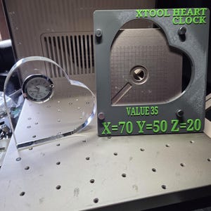 May include: A clear acrylic heart-shaped clock with a silver-rimmed face and a gray rectangular frame featuring the green text XTOOL HEART CLOCK, VALUE 35, and X=70 Y=50 Z=20. The frame contains a metallic heart-shaped insert with a grid pattern.