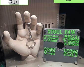 Könnte beinhalten: Eine graue physische Vorrichtung mit der Aufschrift XTOOL PAW und grünem Text, der die Abmessungen für Zylinder-, Rechteck- und Kordeleinstellungen angibt. Daneben eine beige Schaufensterpuppenhand mit einem klaren, sternförmigen Schlüsselanhänger.