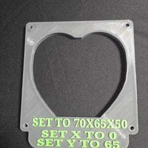 Könnte beinhalten: Ein durchscheinender grauer Kunststoffrahmen mit herzförmigem Ausschnitt. Der Rahmen hat abgerundete Ecken und mehrere kleine Löcher. Der Text "SET TO 70X65X50, SET X TO 0, SET Y TO 65" ist grün gedruckt.
