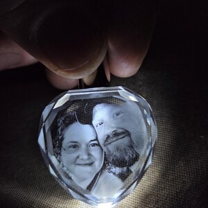 Op de afbeelding: Een hartvormige, heldere kristallen hanger met een lasergegraveerd monochroom portret van twee personen. Het kristal heeft gefacetteerde randen die het licht opvangen, wat een gloeiend effect cre&euml;ert rond de gedetailleerde grijswaardenafbeelding.
