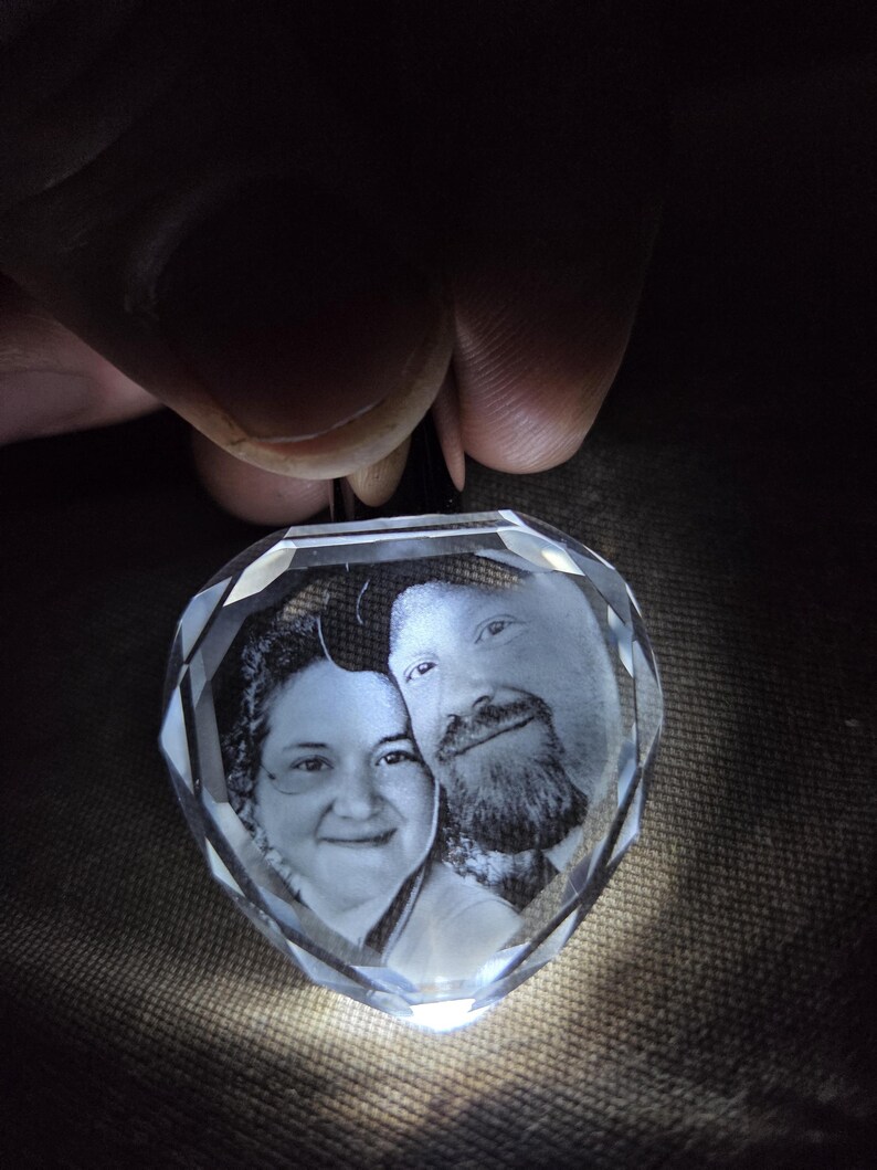 Op de afbeelding: Een hartvormige, heldere kristallen hanger met een lasergegraveerd monochroom portret van twee personen. Het kristal heeft gefacetteerde randen die het licht opvangen, wat een gloeiend effect cre&euml;ert rond de gedetailleerde grijswaardenafbeelding.