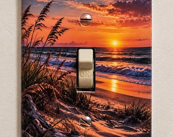 Könnte beinhalten: Eine rechteckige Lichtschalterabdeckung mit einem aufgedruckten Bild eines Strandes bei Sonnenuntergang, orangefarbenen und violetten Wolken, Strandhafer, Treibholz und Meereswellen. Der beige Kippschalter in der Mitte trägt das Wort ON.
