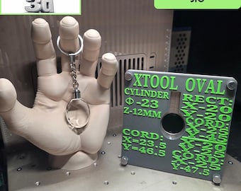 Könnte beinhalten: Ein beiges, 3D-gedrucktes Handmodell, das einen klaren Kristall-Schlüsselanhänger hält, neben einer grauen, quadratischen physischen Vorrichtung mit grünem Text, der XTOOL OVAL, CYLINDER, RECT und verschiedene Koordinatenmaße in Millimetern anzeigt.