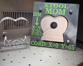 Könnte beinhalten: Ein klares, herzförmiges Acrylobjekt und ein quadratischer, grauer 3D-gedruckter Rahmen mit grünem Text XTOOL MOM, DIM: X=70 Y=55 Z=23.5 und CORD: X=0 Y=65. Der Rahmen hat einen herzförmigen Ausschnitt in der Mitte und vier kleine Metallbefestigungen.