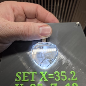 Op de afbeelding: Een grijs rechthoekig apparaat met groene tekst die luidt "SET X=35.2, Y=35, Z=12". Een helder, hartvormig kristal wordt boven de tekst verlicht. Het apparaat wordt door een hand vastgehouden.