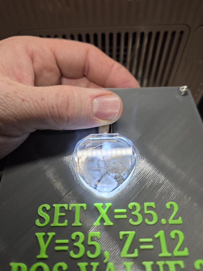 Op de afbeelding: Een grijs rechthoekig apparaat met groene tekst die luidt "SET X=35.2, Y=35, Z=12". Een helder, hartvormig kristal wordt boven de tekst verlicht. Het apparaat wordt door een hand vastgehouden.
