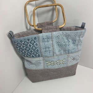 May include: A handbag featuring a patchwork design of light blue denim and solid brown fabric. The bag has two square wooden handles and is decorated with white and dark blue geometric embroidery patterns across the denim sections.