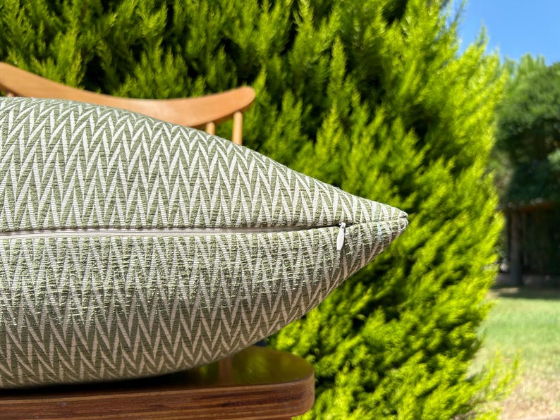 May include: A green and white patterned throw pillow with a zipper closure. The pillow is sitting on a wooden chair.