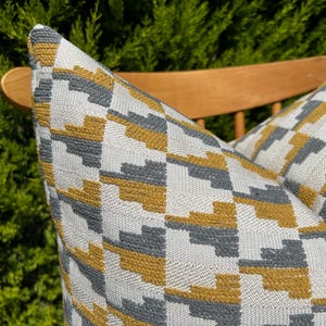 Peut inclure: Deux coussins décoratifs avec un motif géométrique dans des tons de gris et jaune moutarde. Les coussins sont sur une chaise en bois.