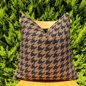 May include: A brown and black patterned throw pillow with a houndstooth design. The pillow is sitting on a wooden chair.
