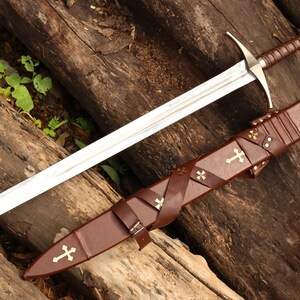 Puede incluir: Una espada de estilo medieval con una hoja plateada y un mango envuelto en cuero marr&oacute;n. La espada descansa sobre una superficie de madera junto a una vaina de cuero marr&oacute;n adornada con cruces doradas. La empu&ntilde;adura de la espada presenta un emblema decorativo.