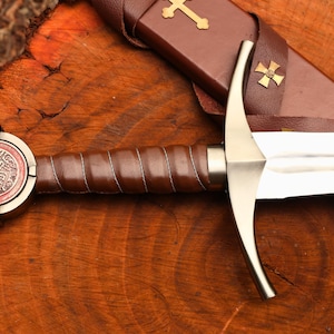 Puede incluir: Una espada de estilo medieval con una hoja y guarda plateadas, y un mango envuelto en cuero marr&oacute;n. El pomo presenta un dise&ntilde;o circular con un emblema rojo y plateado. Se ve una vaina de cuero marr&oacute;n con una cruz dorada.