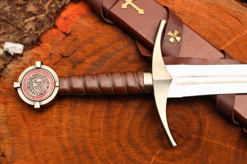 Puede incluir: Una espada de estilo medieval con una hoja y guarda plateadas, y un mango envuelto en cuero marr&oacute;n. El pomo presenta un dise&ntilde;o circular con un emblema rojo y plateado. Se ve una vaina de cuero marr&oacute;n con una cruz dorada.