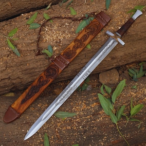 May include: A Viking sword with a silver blade and a brown handle is displayed next to its brown leather sheath. The sheath features carved Celtic knot designs. The blade has etched patterns and text. The sword rests on a wooden surface.