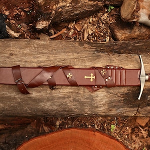 Puede incluir: Una espada medieval con una vaina y empu&ntilde;adura de cuero marr&oacute;n, sobre una superficie de madera. La vaina presenta cruces doradas y correas de cuero. La espada tiene una hoja plateada y una guarda detallada.